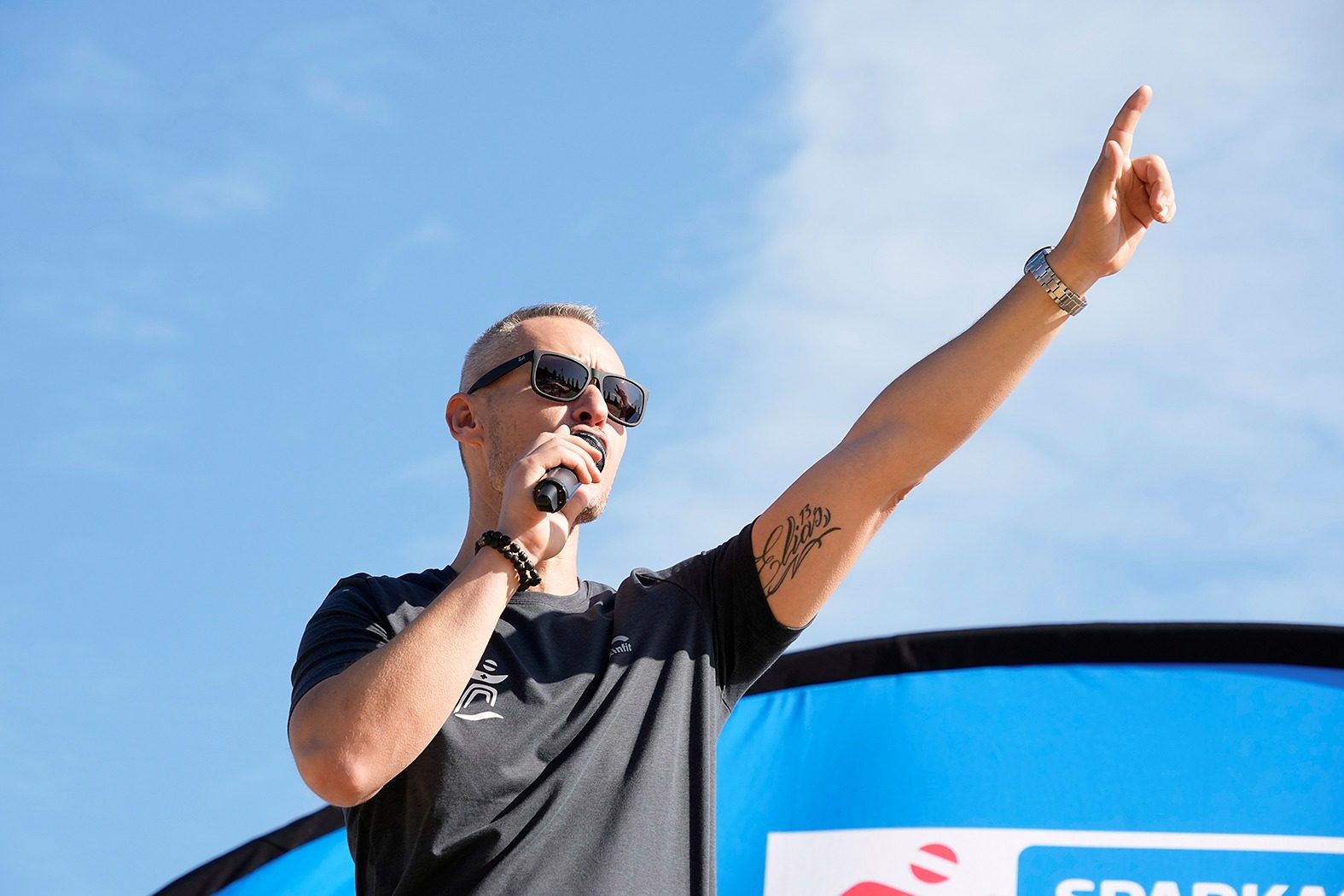 Mike Metelko moderiert bei einem Sportevent im Freien, Mikrofon in der Hand, Arm in den Himmel gestreckt