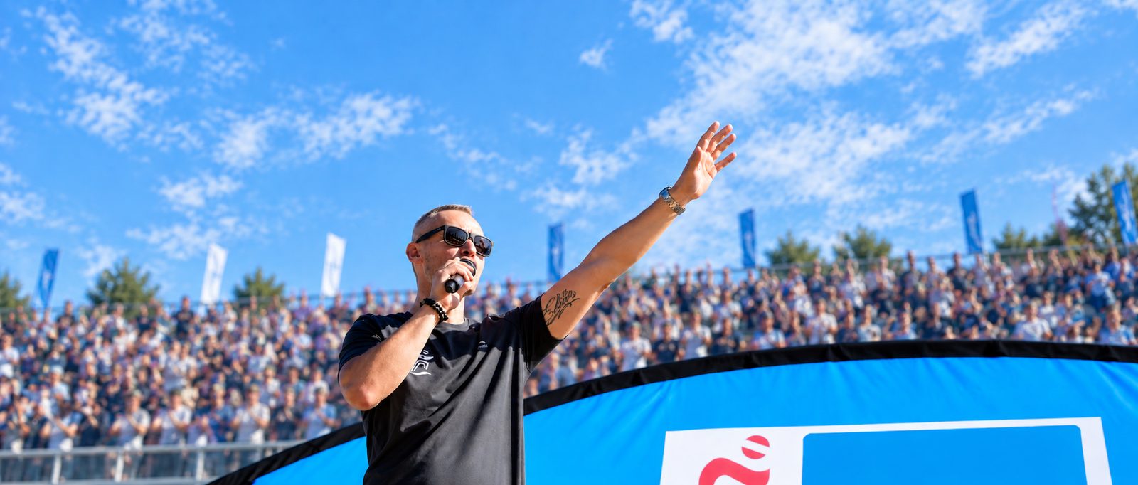 Mike Metelko moderiert ein Sportevent, Mikrofon in der Hand, Arm zum Publikum ausgestreckt
